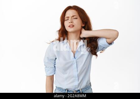 Junge Bürofrau mit Nackenschmerzen, massieren, sich nach hinten reiben und vor Unwohlsein grimacing, stehend in Bluse vor weißem Hintergrund Stockfoto