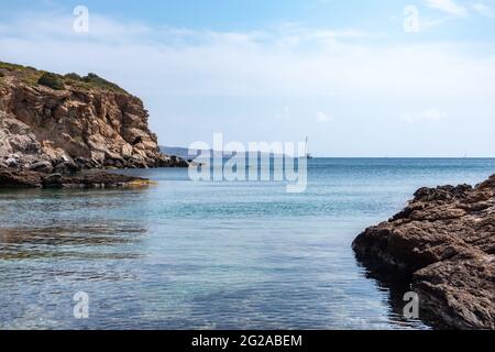 Wildes mittelmeer mit felsigen Klippen, Segelboot in der Ferne und blauem klarem Wasser. Reisen Sie nach Griechenland in der Nähe von Athen. Sommer Natur szenische Lagune Stockfoto