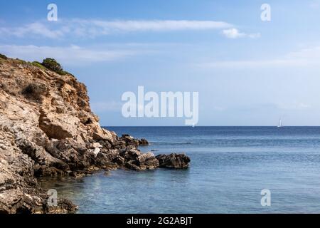Wildes mittelmeer mit felsigen Klippen, Segelboot in der Ferne und blauem klarem Wasser. Reisen Sie nach Griechenland in der Nähe von Athen. Sommer Natur szenische Aussicht Stockfoto