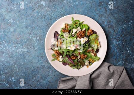 Draufsicht auf gesunde und leckere vegetarische Speisen. Salat mit einer Mischung aus frischen grünen Blättern, Blauschimmelkäse, Nüssen und Senfsoße auf blauem Betonboden Stockfoto