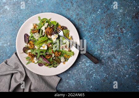 Draufsicht auf gesunde und leckere vegetarische Speisen. Salat mit einer Mischung aus frischen grünen Blättern, Blauschimmelkäse, Nüssen und Senfsoße auf blauem Betonboden Stockfoto