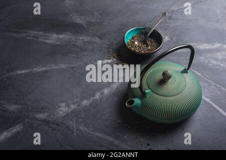 Traditioneller grüner oder türkisfarbener asiatischer gusseiserner Wasserkocher und trockene Teeblätter auf schwarzem Marmorhintergrund mit Kopierfläche Stockfoto