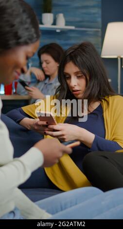 Eine Gruppe gemischter Renner, die spät in der Nacht im Wohnzimmer zusammenhängen und online Nachrichten auf dem Handy suchen. Multirassische Freunde, die während einer Überraschungsparty einen Unterhaltungsfilm ansehen Stockfoto