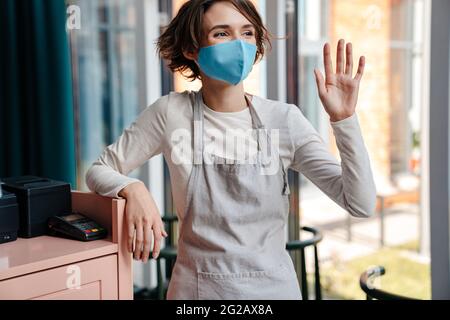 Glückliche weibliche Cafe-Besitzerin trägt eine schützende Gesichtsmaske und öffnet sich nach der Coronavirus-Epidemie wieder Stockfoto