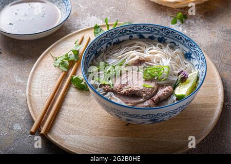 Vietnamesische Nudelsuppe Pho bo in einer Schüssel, garniert mit Basilikum, Minze, Limette, auf betontem Hintergrund. Stockfoto