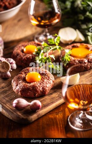 Wunderschön zubereitete Rindersteaks Tartare mit appetitlichen Eigelb-Eiern. Serviert mit Knoblauch, Zwiebeln, Kräutern und Alkohol als Getränk. Weiße Schüssel voller roher Zutaten Stockfoto