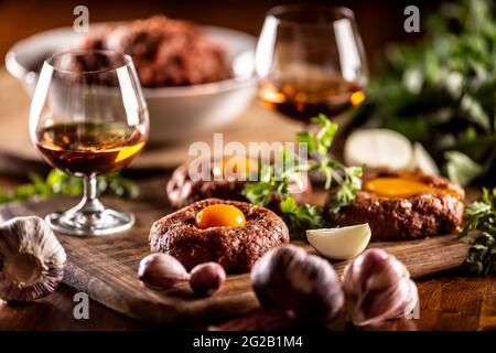 Herzhafte Rindersteak-Tartare werden mit Eigelb, Knoblauch und Kräutern auf einem Holzschreibtisch mit einer weißen Schüssel voller rohem Fleisch im Hintergrund serviert. Whiskey als bev Stockfoto