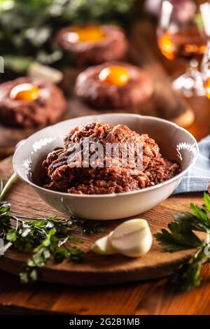 Weiße Schüssel mit rohem Fleisch gefüllt, bereit, köstliche Steak Tartare mit herzhaften Eigelb zuzubereiten. Bereits vorbereitete Tartares im Hintergrund mit Rum und Stockfoto