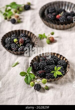 Reife Brombeeren mit Blättern in rustikalen, herben Backformen auf einer Leinentischdecke aus Naturstoff. Stockfoto