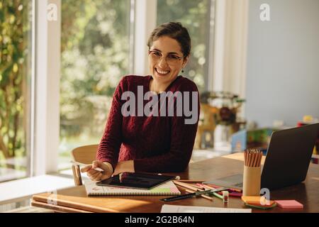 Porträt einer lächelnden Designerin, die zu Hause sitzt. Frau, die mit einem digitalen Tablet an der Illustration arbeitet. Stockfoto