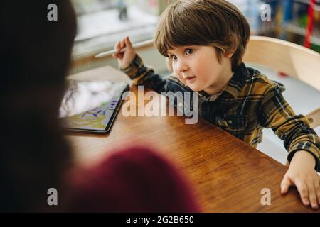 Unschuldiger Junge, der ihre Mutter ansieht, während er mit einem digitalen Tablet am Tisch sitzt. Netter Sohn, der ihre Mutter ansieht, während er auf einem Tablet-pc zeichnet. Stockfoto