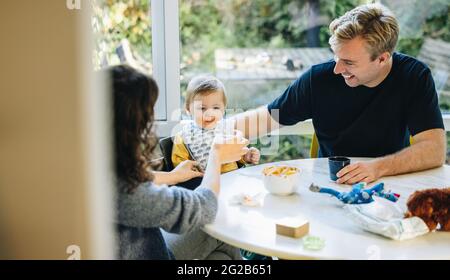 Eltern füttern Baby zu Hause. Mutter und Vater sitzen am Tisch und füttern ihr Kind. Stockfoto