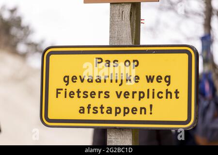 BERGEN AAN ZEE, NIEDERLANDE - 30. Apr 2021: Melden Sie sich in der niederländischen Küstendünenlandschaft an. ÜBERSETZUNG: 'WATCH OUT GEFÄHRLICHE STRASSE. RADFAHRER VERPFLICHTET, ZU DISM Stockfoto