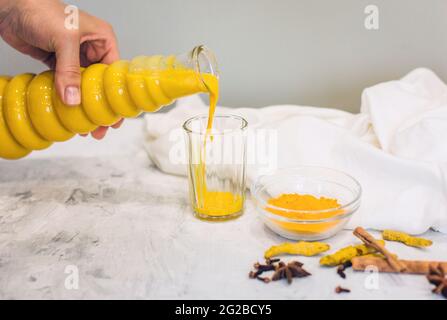 Eine Frauenhand gießt einen Kurkuma-Latte mit Milch und Zimt aus einer Flasche in ein Glas. Ein Elixier der Gesundheit und Kraft. Eine traditionelle gesunde indische d Stockfoto