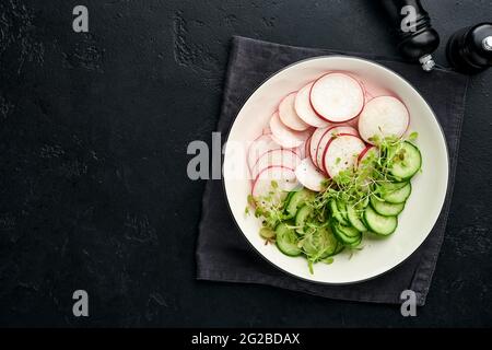 Frischer Salat mit rotem Rettich, Gurke, Gemüse, mikrogrünen Radieschen auf weißem Teller auf grauem Steingrund. Blick von oben. Konzept vegan und hea Stockfoto