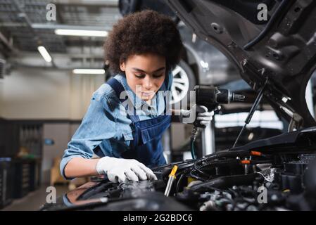 Junger afroamerikanischer Mechaniker in Overalls, der Elektroschrauber hält und mit dem Automotor in der Garage arbeitet Stockfoto