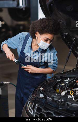 Fokussierter junger afroamerikanischer Mechaniker in Schutzmaske, der Schlüssel hält und mit dem Automotor in der Garage arbeitet Stockfoto