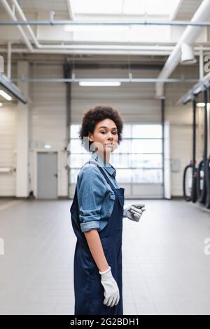 Selbstbewusster junger afroamerikanischer Mechaniker in Handschuhen, der in der Garage auf die Kamera schaut Stockfoto