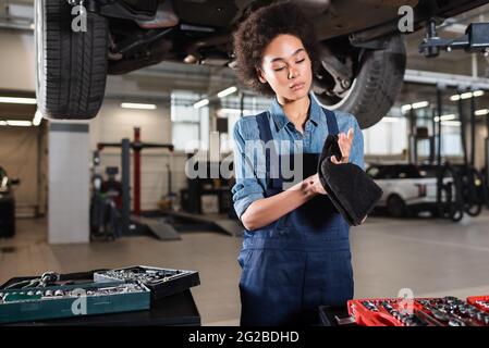 Fokussierter junger afroamerikanischer Mechaniker, der unter dem Auto steht und in der Garage die Hände mit einem Handtuch trocknet Stockfoto