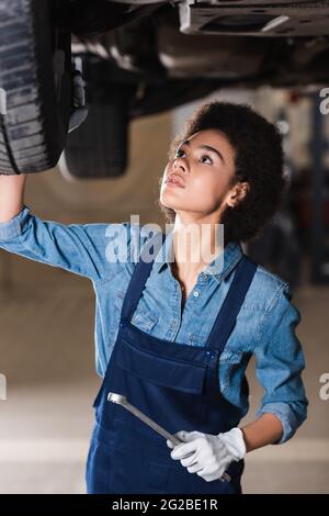 Junger afroamerikanischer Mechaniker mit Schraubenschlüssel in der Hand, der den Boden des Autos in der Garage repariert Stockfoto