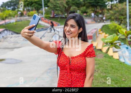 Brünette Frau trägt rotes Kleid mit Smartphone und macht ein Selfie im Skatepark, lächelnd Stockfoto