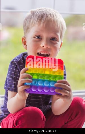 Ein kleiner blonder Junge sitzt und schaut mit einem modernen bunten und hellen popit Spielzeug Simple Grübchen auf die Kamera. Das Kind mit bunten trendigen Antistress s Stockfoto