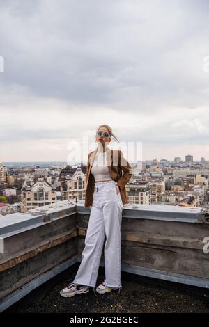 In voller Länge ein junges Modell in blauer Sonnenbrille, das mit der Hand in der Tasche auf dem Dach mit Stadtbild und verschwommenem Vordergrund posiert Stockfoto