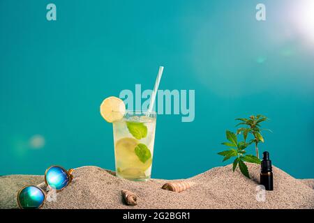 Limonade in verschiedenen Gläsern/Gläsern, CBD-Tropferöl und ein Zweig Marihuanapflanze im Sand. Sonnenbrillen und Muscheln sind von der Seite zu sehen. T Stockfoto