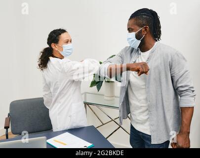 Der afroamerikanische Mann und sein Arzt tragen medizinische Masken und grüßen sich gegenseitig durch Ellenbogenanstiege in der Arztpraxis. Ellenbogenstoß, Kampf Coronavirus epid Stockfoto