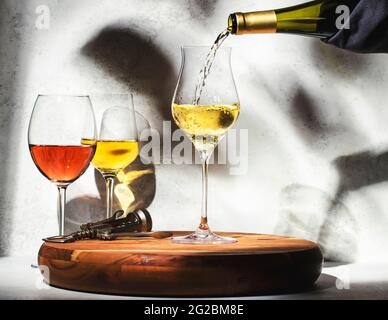 Weinprobe. Weißwein, der auf dem Hintergrund in Glas gegossen wird, mit einer Auswahl an Rot-, Weiß- und Roséweinen in Gläsern. Hartes Sonnenlicht und Schatten von Folia Stockfoto