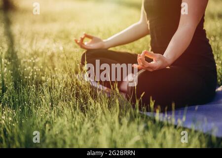 Die junge Kaukasierin praktiziert Yoga in Lotusposition an einem frühen sonnigen Morgen in einem Wald mit Gras und Tau. Gesundes Lifestyle-Konzept. Morgen ich Stockfoto