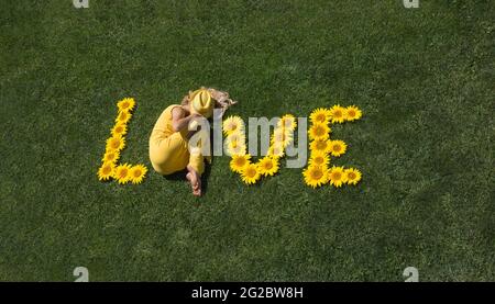 Wortliebe aus Sonnenblumenblumen auf grünem Rasen gelegt. Anstelle des Buchstabens O gibt es ein Mädchen in gelber Kleidung. Sommerstimmung, Blumenfantasien, romanc Stockfoto