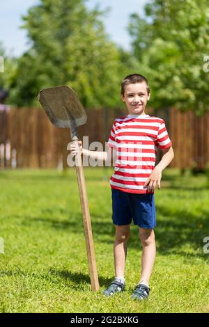 Ein kleiner fröhlicher Junge steht und hält eine Schaufel in der Hand, bereit, im Garten eines Landhauses zu arbeiten. Stockfoto