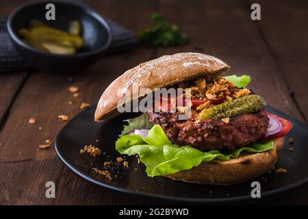 Burger mit vegetarischem Hamburger-Patty, Salat, Tomaten und gerösteten Zwiebeln auf einem schwarzen Teller auf einem Holztisch Stockfoto