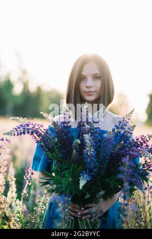 Junge schöne Frau in einem Kleid steht in einem Feld von Lupinen. Mädchen hält einen großen Strauß lila Lupinen in einem blühenden Feld. Blühende Lupinenblüte Stockfoto