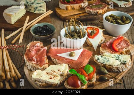 Baguette-Brotscheiben mit Käse, Speck, Salami, Oliven und Kapern als Vorspeise, Tapas, Antipasti, mediterraner Stil auf einem rustikalen Holztisch Stockfoto