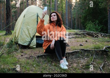 Das Mädchen sitzt neben dem Zelt und fängt das Signal des Mobilfunknetzes im Wald ein. Reisen Sie außerhalb der Stadt im Wald. Camping. Stockfoto