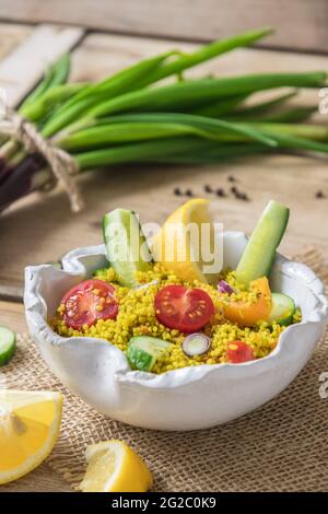 Couscous-Salat mit Gurken, Tomaten und Zwiebeln in einer weißen Schüssel auf rustikalem Holzhintergrund, vertikal Stockfoto