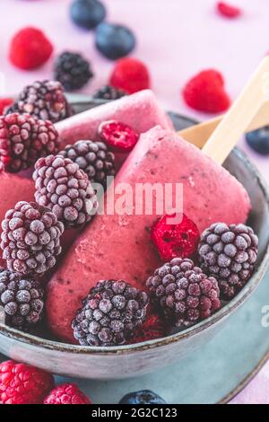 Hausgemachte gemischte Beerenjoghurt-Popsicles in einer Schüssel auf rosa Hintergrund, vertikale Nahaufnahme Stockfoto