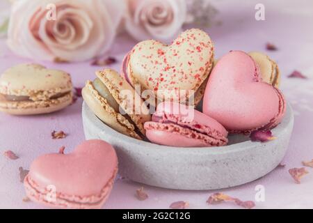 Herzförmige süße Macarons und Rosen in Pastellfarben auf rosa Basis. Ein köstliches Dessert oder Geschenk zum Valentinstag oder Muttertag. Stockfoto