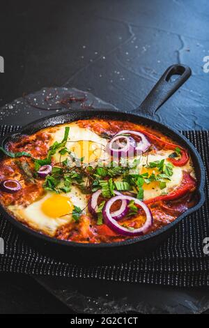 Shakshuka in einer Eisenpfanne. Traditionelles Gericht aus dem Nahen Osten. Spiegeleier mit Tomaten, Paprika, Gemüse und Kräutern, Spiegeleier in sonniger Lage. Vertikal w Stockfoto