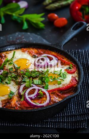 Shakshuka in einer Eisenpfanne. Traditionelles Gericht aus dem Nahen Osten. Spiegeleier mit Tomaten, Paprika, Gemüse und Kräutern, Spiegeleier in sonniger Lage. Vertikal. Stockfoto