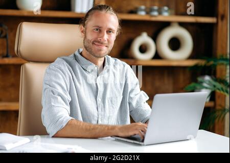 Porträt eines gutaussehenden, selbstbewussten jungen kaukasischen Mannes, CEO, Manager oder Anwalt, der in einem Büro sitzt, ein stilvolles Hemd trägt, einen Laptop benutzt, online arbeitet, die Kamera anschaut, Lächelt freundlich Stockfoto