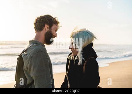 Romantisches glückliches Paar in der Liebe, das am wilden Strand an der Küste steht - Freund und Freundin tragen coole Kleidung und Vintage-Rucksack, der ich stehe Stockfoto