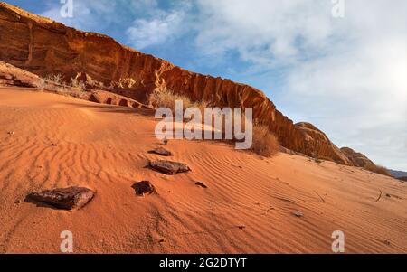 Felsformationen in der Wadi Rum Wüste, helle Sonne scheint auf rotem Sand, Felsen, einige trockene Büsche, blauer Himmel darüber Stockfoto