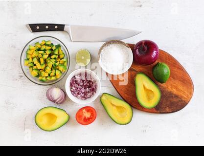 Avocado-Hälften, Limetten und Zwiebeln - Grundzutaten der Guacamole auf weißem Arbeitsbrett, Messer oben, flach liegend Foto Stockfoto