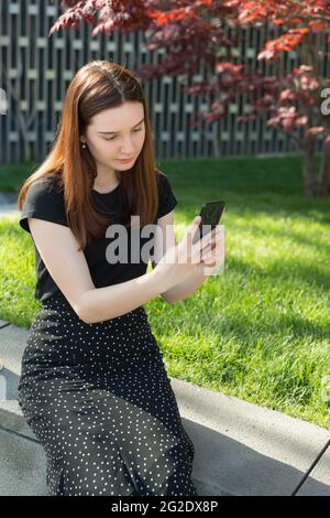 Ein junges, schönes Mädchen mit langen Haaren und schwarzem Sweatshirt und Rock sitzt auf der Straße mit Smartphone in den Händen und schaut auf den Gesprächspartner Stockfoto