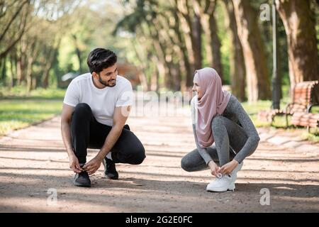 Glücklicher arabischer Mann und Frau, die Schnürsenkel an Sport-Sneakers binden, lächeln und einander im grünen Park anblicken. Konzept eines aktiven und gesunden Lebensstils. Stockfoto