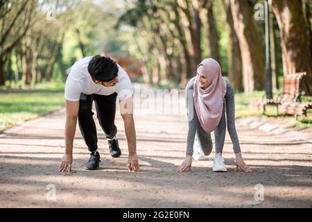 Junges muslimisches Paar in Sportkleidung, das in Startposition für den Lauf im grünen Sommerpark steht. Mann und Frau in Hijab, die sich für das morgendliche Training im Freien vorbereiten. Stockfoto