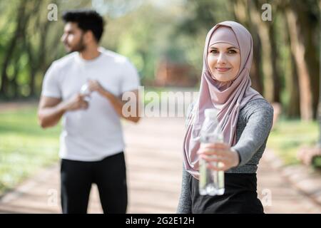 Hübsche Frau im Hijab, die Wasser aus der Flasche hält, während ein muslimischer Mann hinter sich steht und zur Seite schaut. Junge Familie im aktiven Ohr, die den Wasserhaushalt nach dem Training im Freien erneuert. Stockfoto